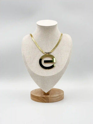 Gold-tone necklace with a broad flat chain and lowercase e-shaped pendant displayed on a fabric jewellery bust.