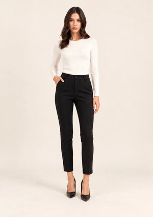 Full-length front view of black tailored ankle-length trousers, worn with an ivory top, with one hand in a pocket.