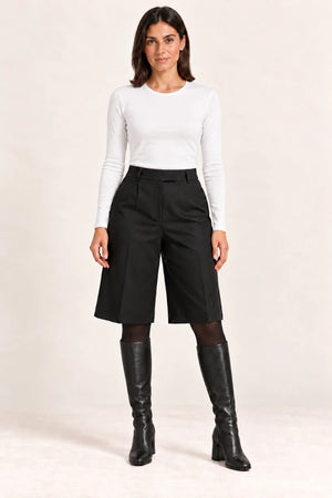 Model wearing Shorts Pinces Nero black pleated shorts with a white top, black tights and knee-high boots