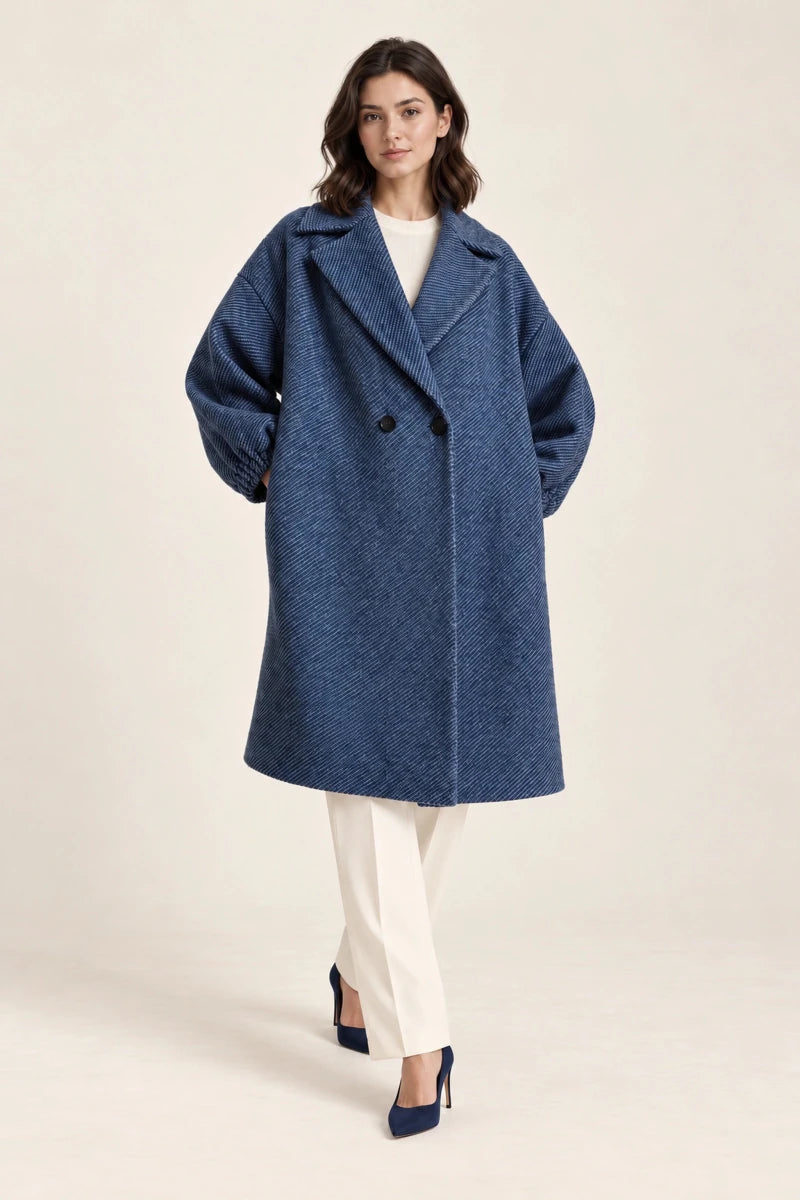 Model stepping forward in Cappotto Oversize Blu/Nero, a blue double-breasted coat with wide lapels, ivory trousers and navy heels.