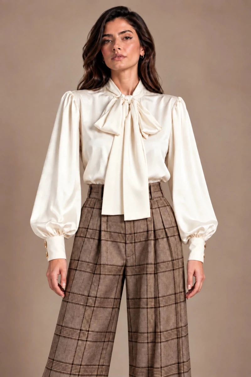 Front view of a model wearing an ivory bow-neck blouse with gathered sleeves and brown checked trousers.