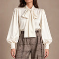 Front view of the Loredana Panna Bow-Neck Shirt in ivory, with a large neck bow and long gathered sleeves, worn with checked trousers.