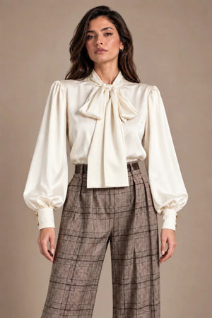 Front view of the Loredana Panna Bow-Neck Shirt in ivory, with a large neck bow and long gathered sleeves, worn with checked trousers.