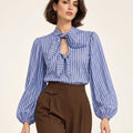 Close-up front view of a blue and brown striped shirt with bow ties and gathered long sleeves, worn with brown trousers.