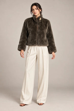Full-length styling view of the brown Mora Moro plush jacket with a raised collar and wide turned-back cuffs, worn with ivory trousers.