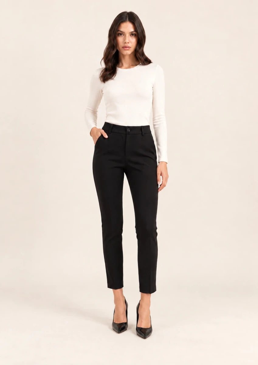 Full-length front view of black tailored ankle-length trousers, worn with an ivory top, with one hand in a pocket.