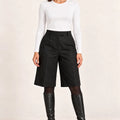 Model wearing Shorts Pinces Nero black pleated shorts with a white top, black tights and knee-high boots