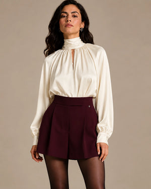 Front styling view of the ivory Zendaya Panna gathered-neck shirt with a high collar and keyhole opening, worn with burgundy shorts.